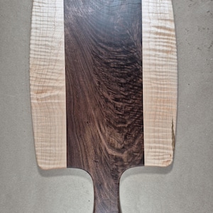 May include: A wooden cutting board with a handle. The board is rectangular with rounded corners. It features a central dark brown stripe flanked by lighter wood on either side. The handle has a hole for hanging.