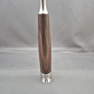 May include: A wooden pepper mill with a dark brown finish and a silver-colored metal top and bottom. The mill has a curved, ergonomic design and a sleek, modern aesthetic. The pepper mill is designed for grinding peppercorns.