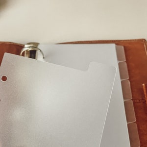 Könnte beinhalten: Ein mattierter Plastik-Teiler für einen Ringordner. Der Teiler hat oben eine Lasche und ist für einen Standard-Ringordner mit 6 Ringen ausgelegt.