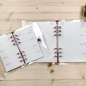 Könnte beinhalten: Zwei transparente Plastikordner mit weißen Seiten und roségoldenen Ringen. Die Ordner sind geöffnet, um die monatlichen Kalenderseiten zu zeigen. Der kleinere Ordner hat den Monat Mai 2021, und der größere Ordner hat die Monate Juni und Juli 2021.