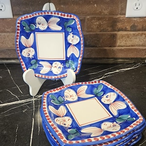 May include: A set of square ceramic plates featuring a central cream-colored square bordered by a blue frame. The border is decorated with painted fish in shades of cream and orange, green foliage, and a red-dotted trim along the scalloped edges.
