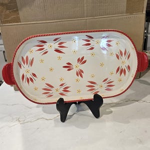 May include: An oval ceramic serving platter with a cream-colored base, featuring a repeating pattern of red leaf-like petals and small yellow dots. The platter has a red rope-textured rim and two red side handles, displayed on a black stand.