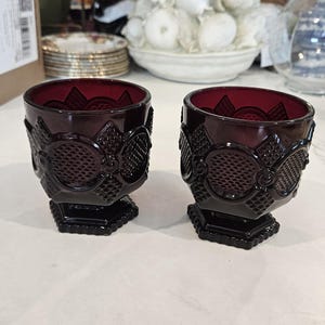 May include: Two deep red glass tumblers featuring a pressed diamond pattern and a circular motif around the exterior. Each glass has a thick, hexagonal base and a wide, open rim, showcasing a dark, translucent colour and a textured surface design.