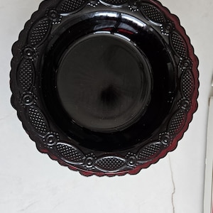 May include: A deep red, circular glass bowl featuring a scalloped edge and a decorative border pattern consisting of alternating diamond-textured ovals and small circular motifs. The center of the bowl is smooth, dark, and reflective.