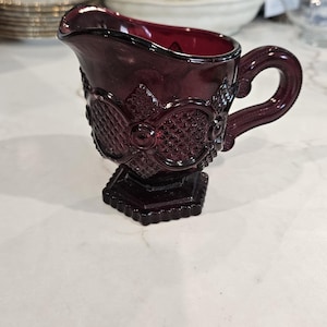 Pode incluir: Uma leiteira de vidro em tom bordô profundo com um padrão de diamante prensado e uma borda recortada. O recipiente possui uma alça curva e assenta sobre uma base hexagonal, exibindo uma cor vermelha escura translúcida e rica.