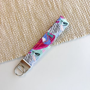 May include: A fabric wristlet keychain featuring a colourful abstract pattern with shades of light blue, magenta, and black and white striped circles. The strap is secured with a silver-toned metal clamp and a circular split ring for keys.
