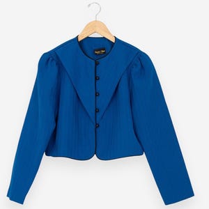 Könnte beinhalten: Eine leuchtend blaue Jacke mit einem einzigartigen Kragendesign und schwarzen Knöpfen. Die Jacke hat lange Ärmel und eine taillierte Passform. Das Kleidungsstück hängt an einem Holzbügel vor weißem Hintergrund. Der Stil der Jacke ist Vintage.