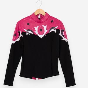 May include: A long-sleeved black top with a high collar and a pink yoke. The yoke is decorated with white and black abstract designs and small black diamond shapes. The top is hanging on a wooden hanger.