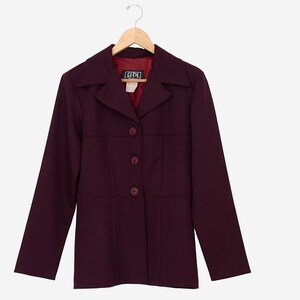 May include: A burgundy blazer with a notched collar and three-button closure. The long-sleeved jacket has a tailored fit and a red interior lining. The blazer is hanging on a wooden hanger.