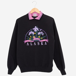 May include: Black sweatshirt with a pink collar and a floral design. The design features purple irises, yellow flowers, and the word "ALASKA" in purple lettering. The sweatshirt is hanging on a wooden hanger.