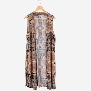 Op de afbeelding: Een mouwloos vest met open voorkant en een patroon in bruin-, zwart- en beigetinten. Het vest hangt aan een houten hanger. Het patroon bevat geometrische en bloemmotieven.