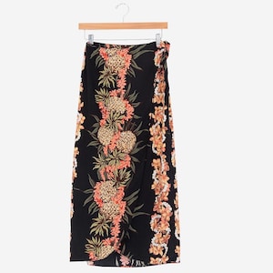 Peut inclure: Jupe portefeuille noire avec imprimé tropical d'ananas et de fleurs. La jupe présente un motif répété d'ananas et de fleurs roses et oranges. La jupe est suspendue à un cintre en bois.