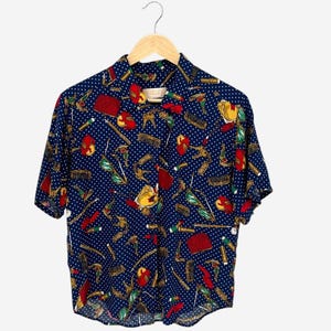 May include: A short-sleeved button-down shirt in dark blue with a white polka dot pattern. The shirt features a colorful print of various small objects including red purses, gold-toned combs, and green accessories scattered across the fabric.