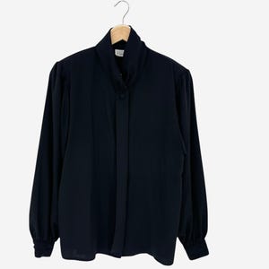May include: A solid navy blue long-sleeved blouse featuring a high standing collar, a concealed button-front placket, and gathered shoulders. The garment has long sleeves with buttoned cuffs and is displayed on a wooden hanger against a plain background.
