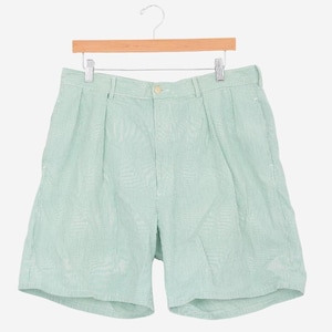 May include: Light green shorts with a subtle striped pattern, hanging on a wooden hanger. The shorts have a button closure and pleats at the front. The fabric appears to be lightweight, suitable for warm weather.