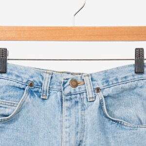 May include: Light blue denim jeans featuring a high-waisted design with a central brass-toned button closure, belt loops, and two front pockets. The denim fabric has a classic light wash finish with visible stitching details throughout the garment.