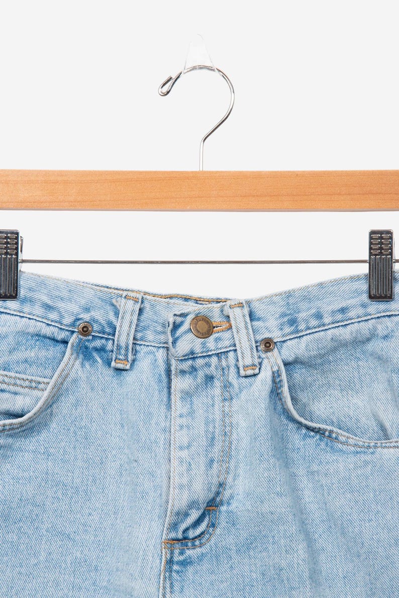May include: Light blue denim jeans featuring a high-waisted design with a central brass-toned button closure, belt loops, and two front pockets. The denim fabric has a classic light wash finish with visible stitching details throughout the garment.