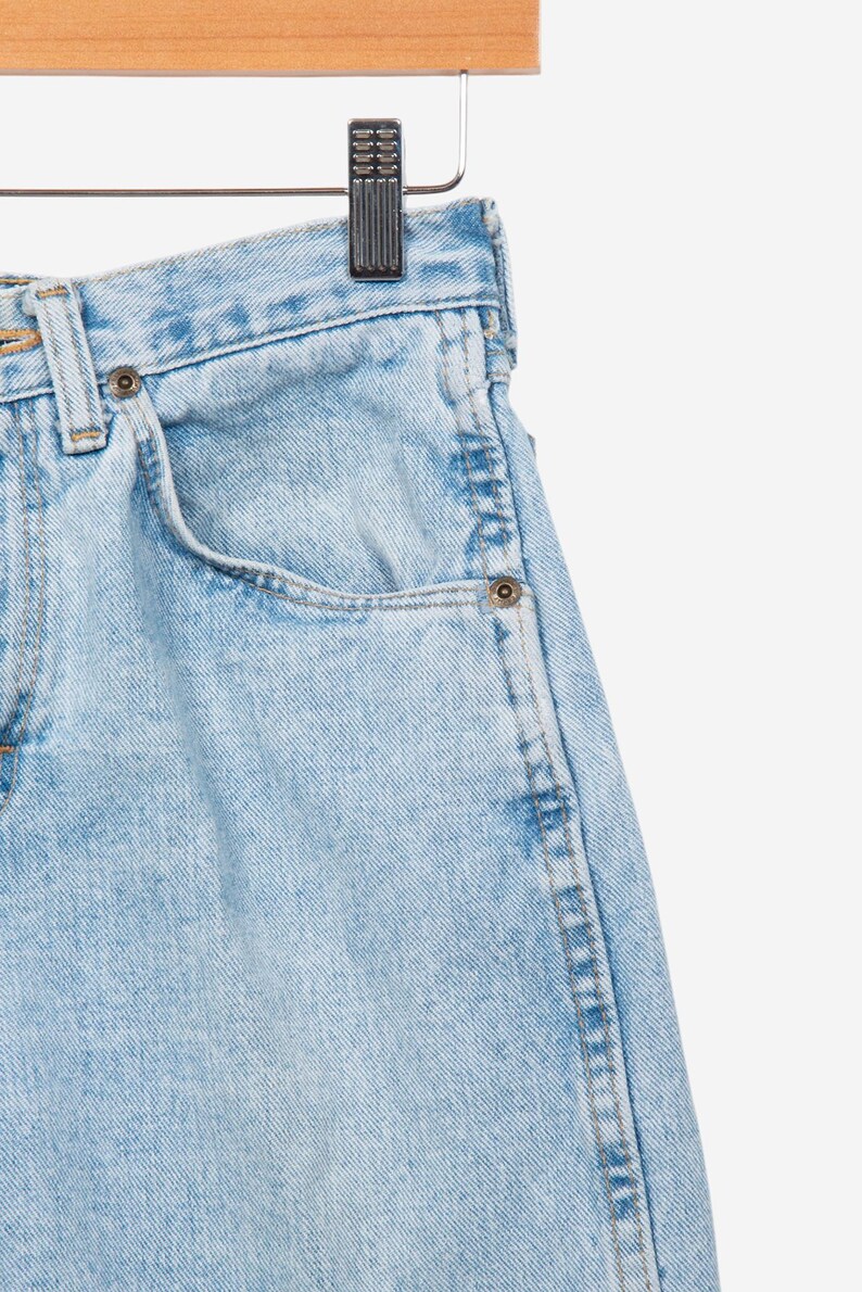 May include: Light blue denim jeans featuring a high-waisted design with a classic five-pocket construction, metal rivets, a button fly closure, and visible stitching details, suspended from a wooden hanger by a metal clip.
