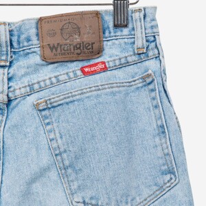 May include: Light blue denim jeans featuring a brown leather-like patch on the waistband with the text Wrangler Premium Quality and a small red rectangular tag on the back pocket with the white text Wrangler stitched in a classic font style.