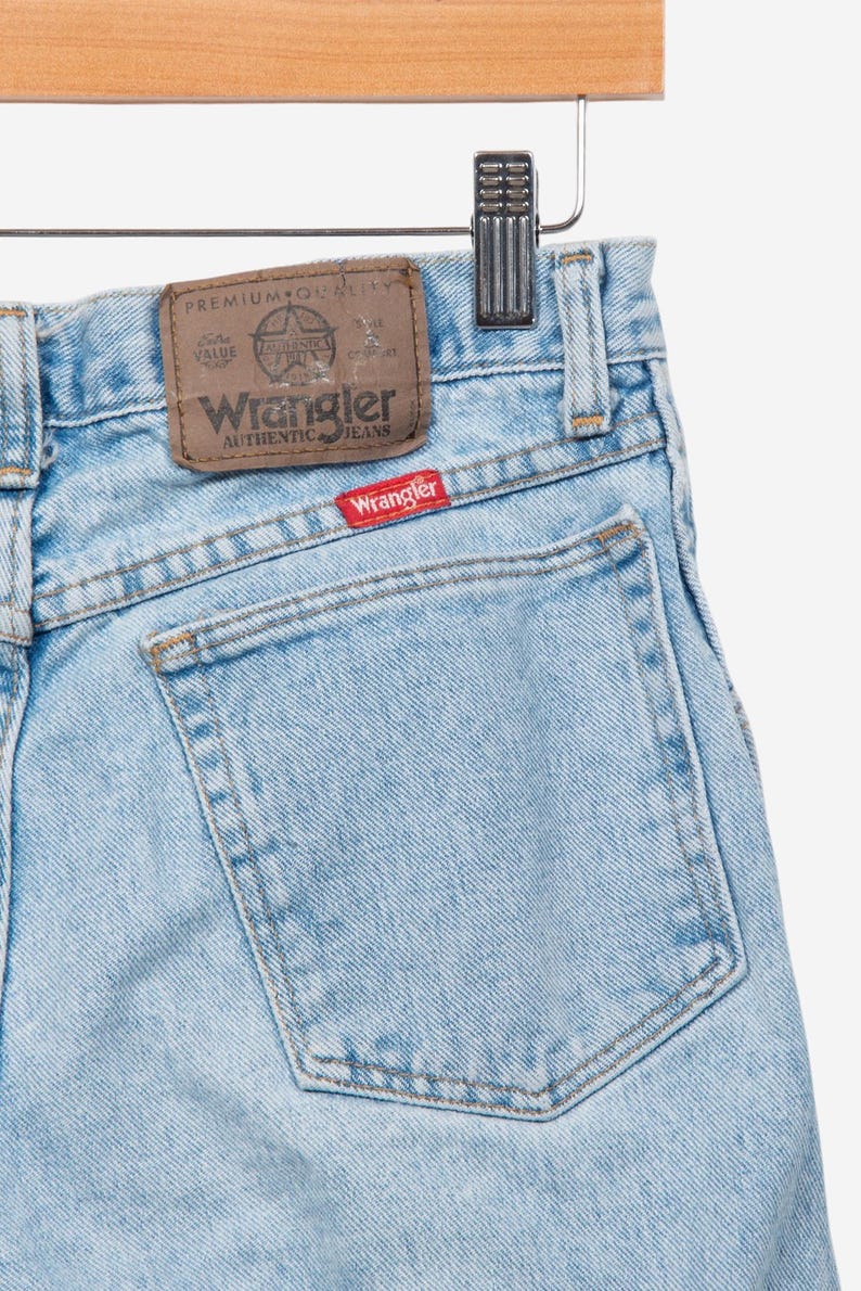 May include: Light blue denim jeans featuring a brown leather-like patch on the waistband with the text Wrangler Premium Quality and a small red rectangular tag on the back pocket with the white text Wrangler stitched in a classic font style.