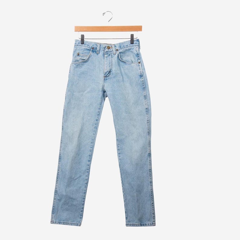 May include: Light blue denim jeans featuring a classic five-pocket design, a button fly closure, and a straight-leg cut. The pants are displayed hanging from a wooden hanger, showcasing a faded wash finish with visible stitching details throughout.