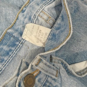 May include: Light blue denim jeans featuring a metal button closure and a white interior tag with the text 96501S, 28 x 30, A829-025, 100% COTTON, and MADE IN USA. The fabric has visible stitching and a classic light wash texture throughout the garment.