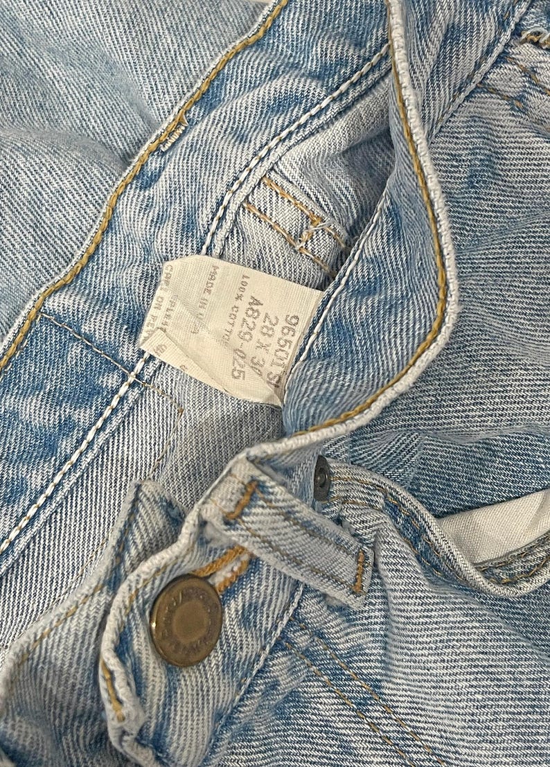 May include: Light blue denim jeans featuring a metal button closure and a white interior tag with the text 96501S, 28 x 30, A829-025, 100% COTTON, and MADE IN USA. The fabric has visible stitching and a classic light wash texture throughout the garment.