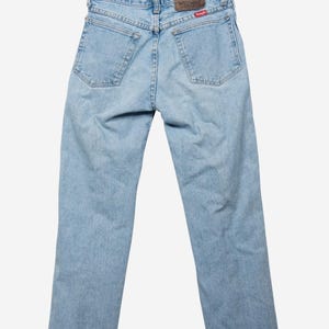 May include: Light blue denim jeans featuring a classic five-pocket design, a brown leather patch on the back waistband with the Wrangler logo, and a small red Wrangler brand tag attached to the right back pocket on a wooden hanger.