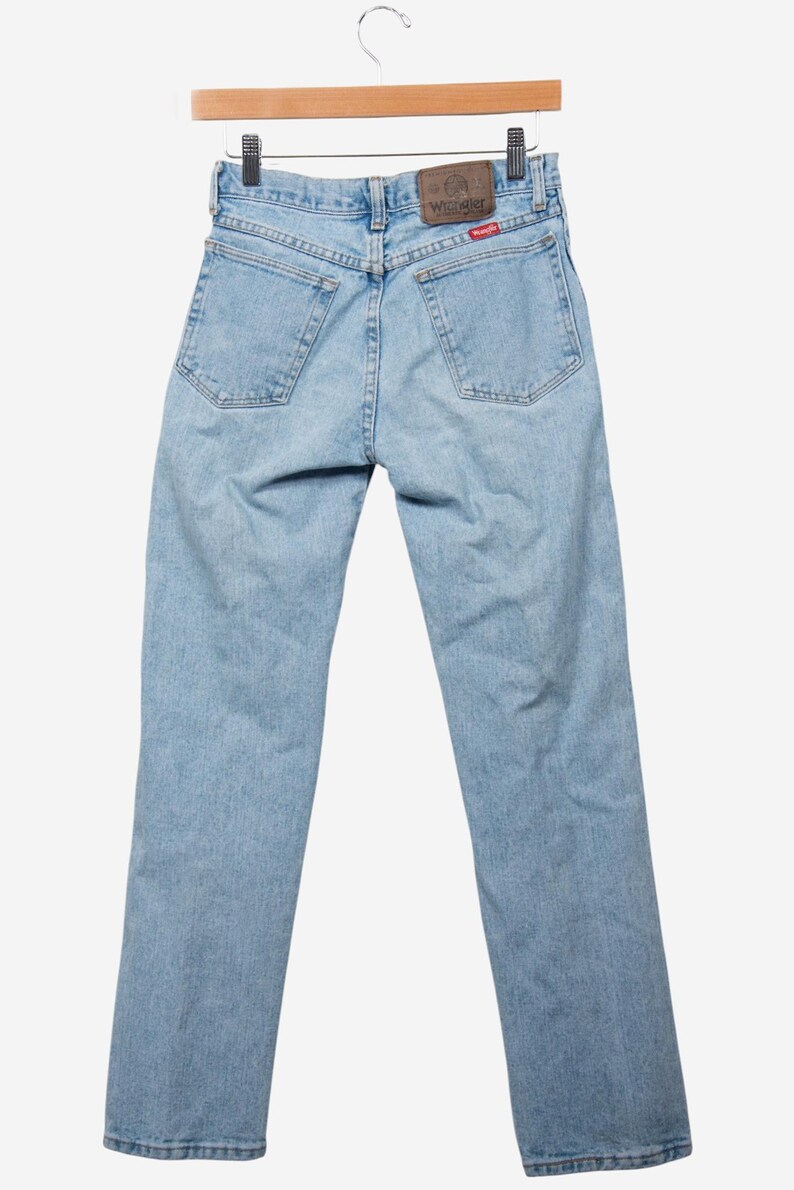 May include: Light blue denim jeans featuring a classic five-pocket design, a brown leather patch on the back waistband with the Wrangler logo, and a small red Wrangler brand tag attached to the right back pocket on a wooden hanger.