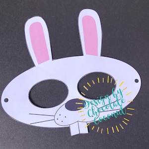 May include: White bunny mask with pink ears, black eye holes, and a black nose. The mask has a white outline of a mouth and whiskers. The text "Design by Chocolate Coconut" is printed on the mask in gold glitter.