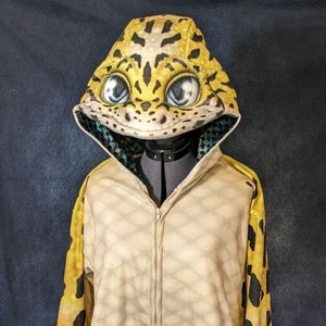 Puede incluir: Un mono beige de cuerpo entero con cremallera y estampado de leopardo amarillo y negro. El mono tiene una capucha con un dise&ntilde;o de cabeza de gecko en 3D.
