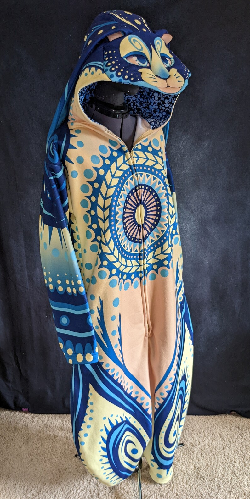 May include: A blue and yellow hooded jumpsuit with a cat face on the hood. The jumpsuit has a mandala pattern on the front and a zipper down the front.