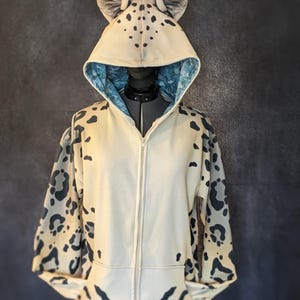 以下が含まれることがあります： クリーム色のジップアップパーカー、ヒョウ柄のデザイン。フードにはグレーの耳と黒い斑点がついています。フードの内側は青と白の模様の裏地が付いています。袖とパーカーの下部にもヒョウ柄があります。