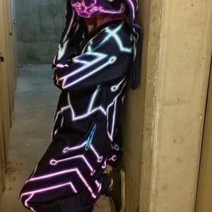 May include: A person wearing a dark blue hooded jumpsuit with a neon pink and blue circuit board design. The hood is shaped like a bunny head.
