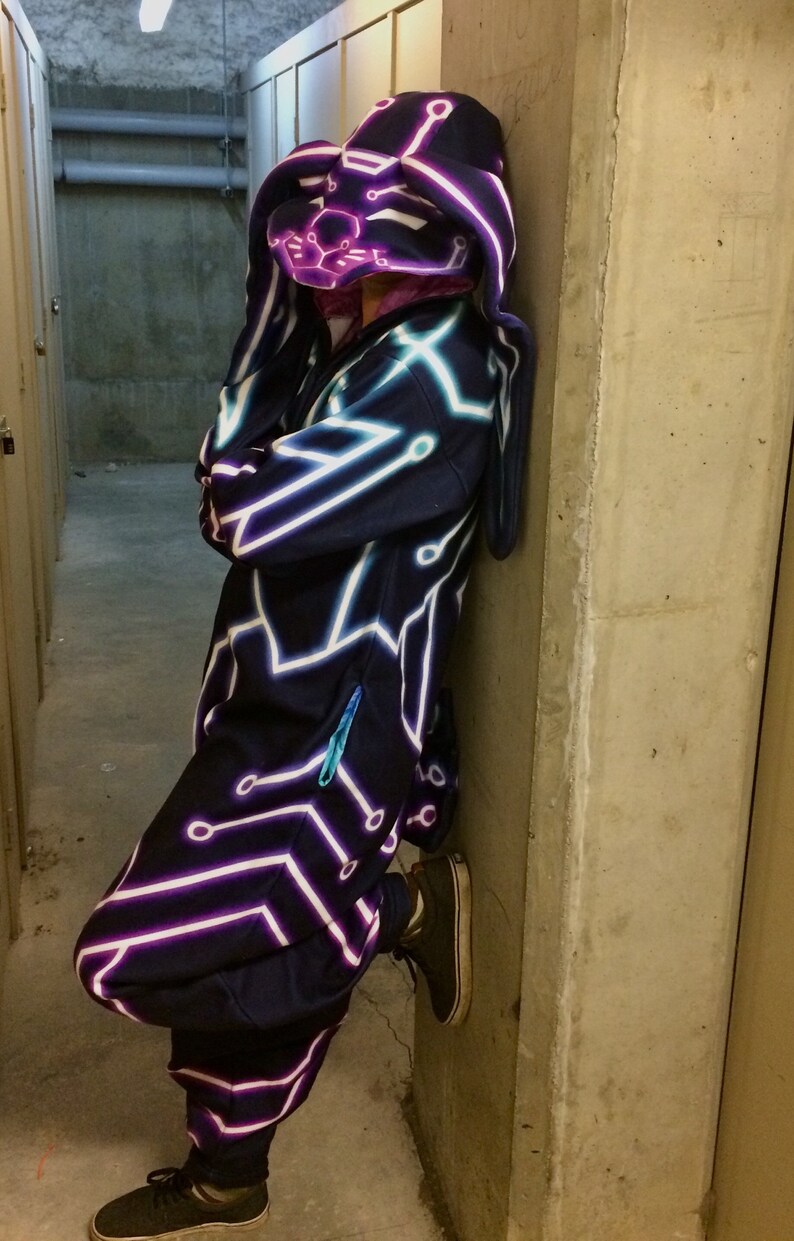 May include: A person wearing a dark blue hooded jumpsuit with a neon pink and blue circuit board design. The hood is shaped like a bunny head.
