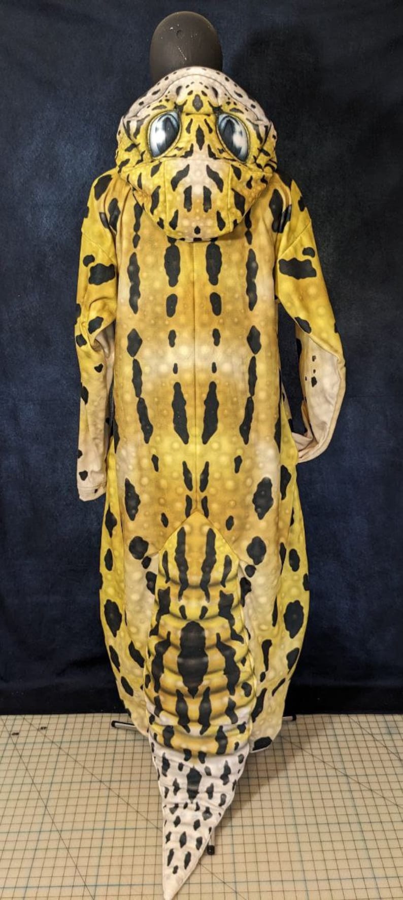 Puede incluir: Un disfraz de gecko leopardo de cuerpo entero con un patr&oacute;n amarillo, negro y blanco. El disfraz tiene una capucha con una cara de gecko realista y una cola larga.