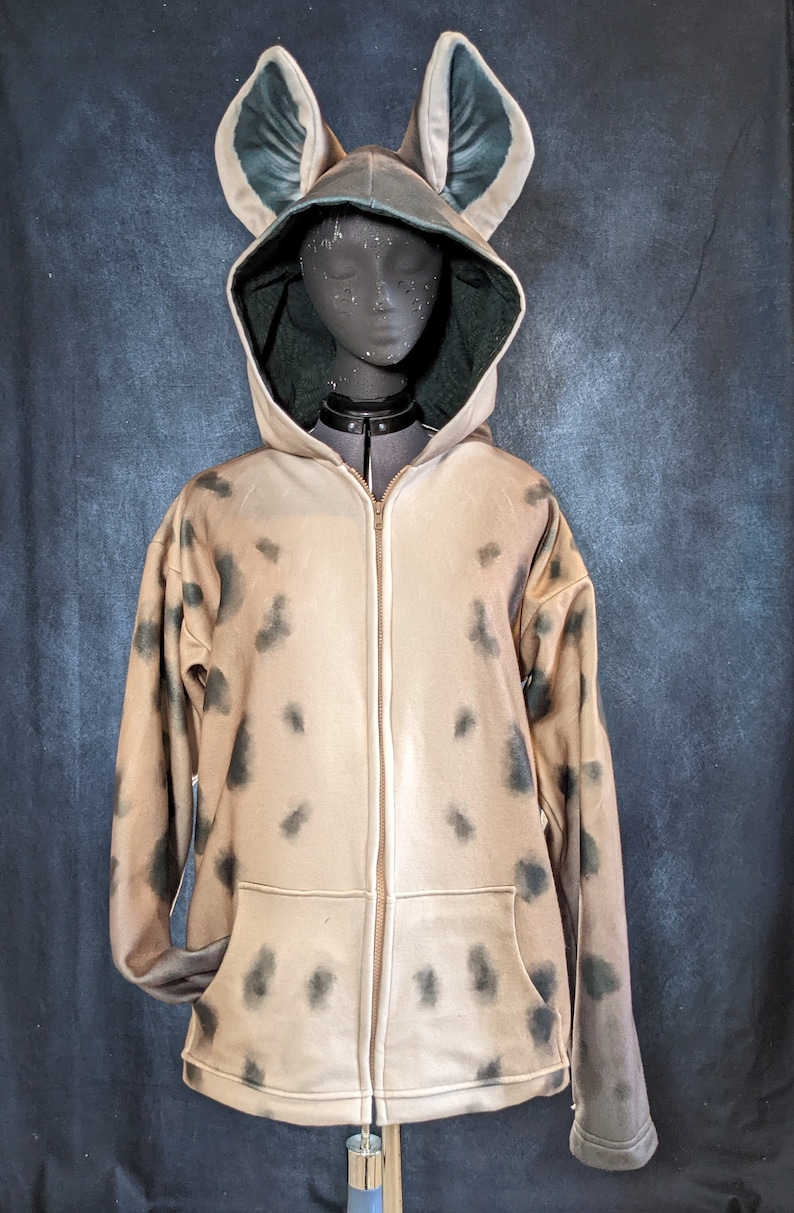 May include: A tan hooded sweatshirt with a zipper closure and black spots. The hood has large, green, pointed ears.