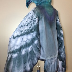 Puede incluir: Una sudadera con capucha azul con un dise&ntilde;o de ala de p&aacute;jaro en las mangas y la espalda. Las alas est&aacute;n detalladas con plumas y tienen un aspecto realista.