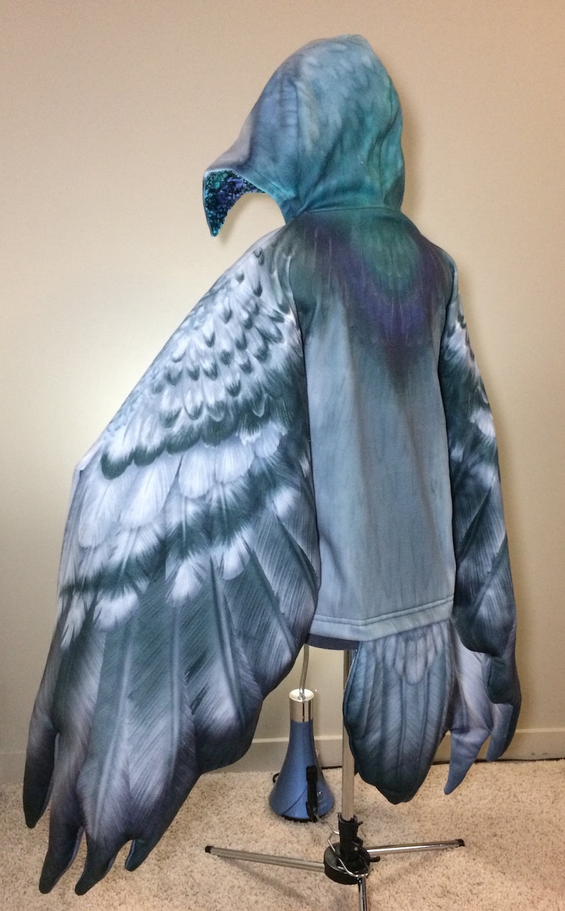 Puede incluir: Una sudadera con capucha azul con un dise&ntilde;o de ala de p&aacute;jaro en las mangas y la espalda. Las alas est&aacute;n detalladas con plumas y tienen un aspecto realista.