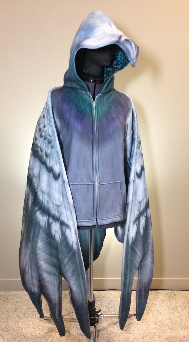 Puede incluir: Una sudadera con capucha gris con cierre de cremallera y alas adjuntas. Las alas est&aacute;n impresas con un patr&oacute;n de plumas realista en tonos grises y azules.