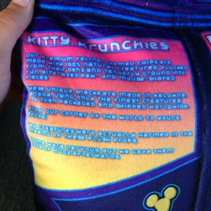 May include: A plush toy shaped like a bag of chips with the text "Kitty Krunchies" and a description of how the chips are made. The bag is blue, purple, yellow, and orange.