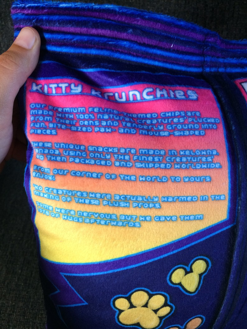 May include: A plush toy shaped like a bag of chips with the text "Kitty Krunchies" and a description of how the chips are made. The bag is blue, purple, yellow, and orange.