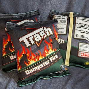 May include: Three black and green chip bags with the words "Trash" and "Dumpster Fire" printed on them. The bags are made of a soft, plush material and are designed to look like real chip bags.