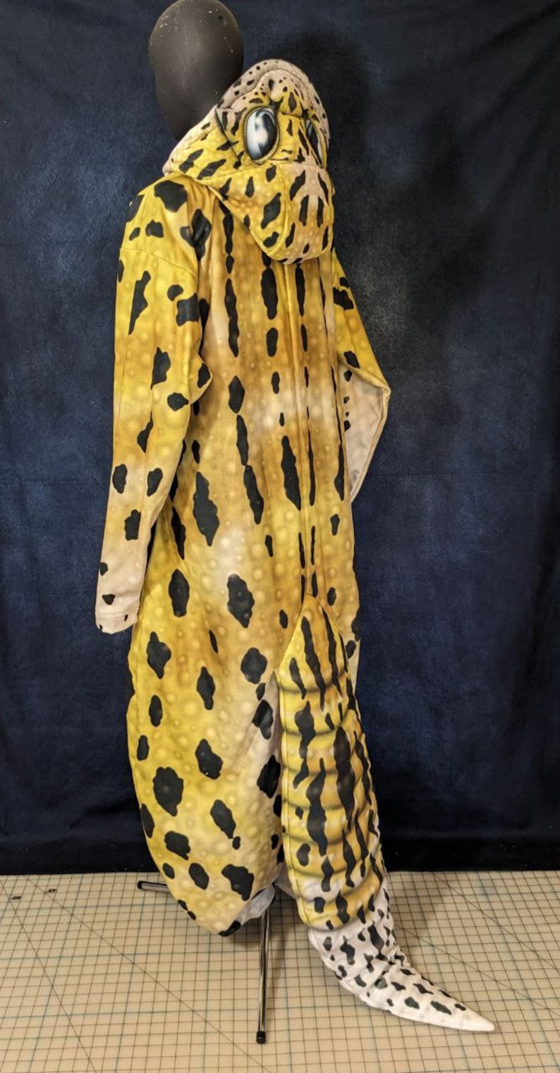 Puede incluir: Un disfraz de animal de cuerpo entero, de color amarillo y negro, con estampado de leopardo y capucha. La capucha tiene una cabeza de leopardo grande, de aspecto realista, de color amarillo y negro, con ojos negros.