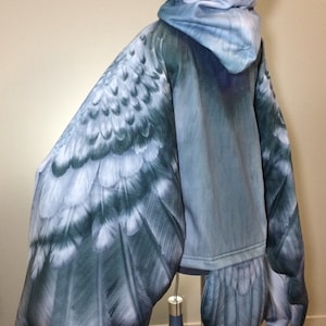 Puede incluir: Una sudadera con capucha azul con un dise&ntilde;o de ala de p&aacute;jaro realista en las mangas y la espalda. Las alas est&aacute;n detalladas con plumas y tienen un degradado de colores azul y gris.