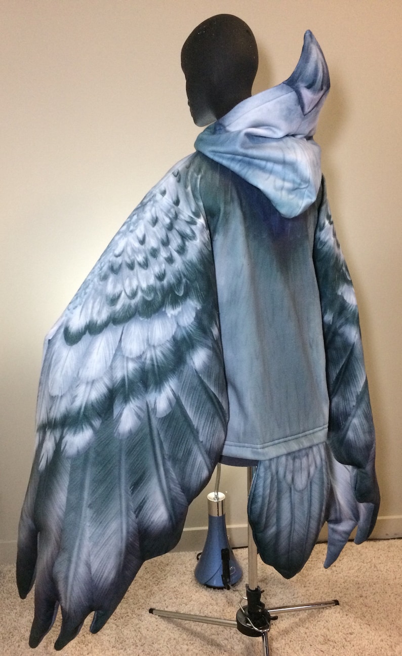 Puede incluir: Una sudadera con capucha azul con un dise&ntilde;o de ala de p&aacute;jaro realista en las mangas y la espalda. Las alas est&aacute;n detalladas con plumas y tienen un degradado de colores azul y gris.