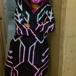 May include: A black hooded jumpsuit with a neon purple and white geometric pattern. The hood has a cat face design with neon purple and white details.