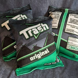 May include: Three green and black plush toys shaped like bags of chips. The bags are labeled "Trash Original" and "Trash". The bags have a green and black design with a white checkmark. The bags are made in Canada.