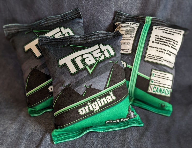 May include: Three green and black plush toys shaped like bags of chips. The bags are labeled "Trash Original" and "Trash". The bags have a green and black design with a white checkmark. The bags are made in Canada.