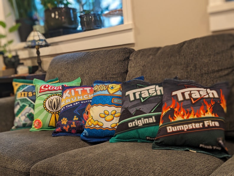 May include: Six colorful throw pillows with different designs and text. The pillows are arranged on a gray couch. The pillows feature designs of a seed packet, a cat food bag, a snack bag, and two different trash bag designs. The text on the pillows includes "Seed", "KITT KRUNCH", "Trash original", and "Trash Dumpster Fire".